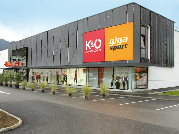 Ein modernes Geschäftshaus mit einem großen rot-orangen Schild mit der Aufschrift „K&O Kastner & Öhler“ und „giga sport“. Die Außenfassade ist mit dunklen Paneelen und großen Schaufenstern versehen, in denen Schaufensterpuppen zu sehen sind. Im Vordergrund ist ein kleiner Parkplatz zu sehen.