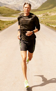 Eine Person läuft auf einer asphaltierten Straße, im Hintergrund sind grüne Hügel und Berge zu sehen. Sie trägt ein schwarzes Langarmshirt, schwarze Shorts und neongrüne Schuhe. Die Sonne wirft einen Schatten auf die Straße.