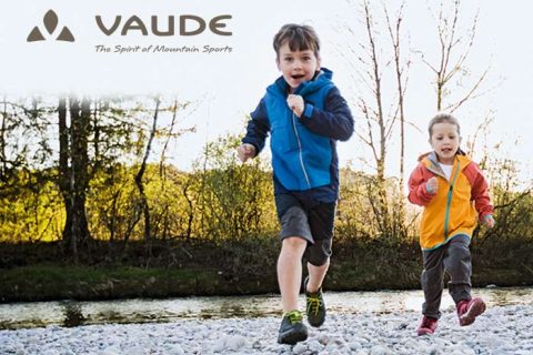 Zwei Kinder in Jacken laufen fröhlich auf einem steinigen Pfad in der Nähe eines Flusses. Das Kind links trägt eine blaue Jacke und das Kind rechts eine gelb-orange Jacke. Im Hintergrund sind Bäume und ein heller Himmel zu sehen. Oben links ist das „VAUDE“-Logo zu sehen.