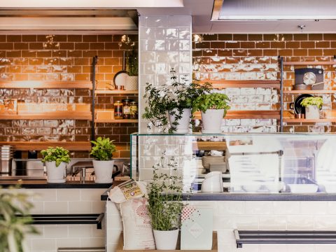 Ein modernes Café-Interieur mit glänzenden braunen U-Bahn-Fliesen an den Wänden. Topfpflanzen und Beutel mit Kaffeebohnen stehen auf Regalen und Theken und sorgen für Grün. Die Atmosphäre ist warm und einladend.