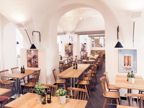 Ein helles Restaurant-Interieur mit in Reihen angeordneten Holztischen und -stühlen. Auf den Tischen stehen Pflanzen und Flaschen, und gerahmte Kunst schmückt die weißen Wände. Von der Decke hängen moderne Pendelleuchten, die eine einladende Atmosphäre schaffen.