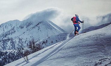 Eine Person in blauer Skiausrüstung und rotem Rucksack fährt einen schneebedeckten Berghang hinauf. Die Szene spielt sich vor einer Kulisse majestätischer, schneebedeckter Gipfel und einem wolkigen Himmel ab.