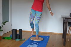 Eine Person in einem roten Hemd und bunten Leggings balanciert auf einem hölzernen Balance Board, das auf einer blauen Trainingsmatte steht. In der Nähe liegen Schaumstoffrollen und Widerstandsbänder auf einem Holzboden.