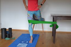 Eine Person in einem roten Hemd und bunten Leggings führt mithilfe eines grünen Widerstandsbands, das an einem Tisch befestigt ist, eine Beinstreckübung durch. Sie steht auf einer blauen Trainingsmatte mit Schaumstoffrollen in der Nähe.