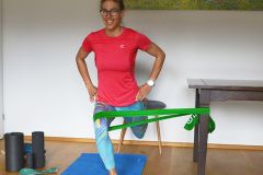 Eine Person in einem roten Hemd und blauen Leggings trainiert mit einem grünen Widerstandsband unter ihrem Knie, das von einem Tisch gestützt wird. Sie lächelt und steht auf einer blauen Matte mit Schaumstoffrollen in der Nähe.