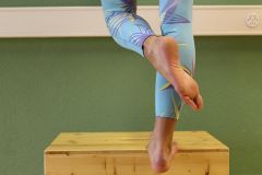 Eine Person in bunten, gemusterten Leggings balanciert auf dem Rand einer Holzkiste vor einer grünen Wand. Nur die Unterschenkel und Füße sind sichtbar, wobei ein Fuß leicht über dem Boden steht.