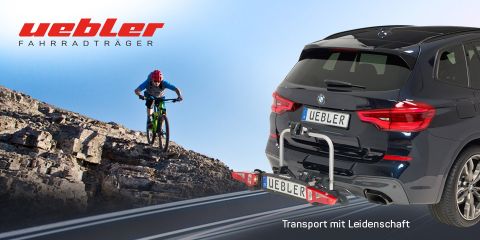 Ein Radfahrer in roter Ausrüstung fährt mit seinem Mountainbike einen steinigen Pfad hinunter. Auf der Straße parkt ein schwarzer SUV mit einem Uebler-Fahrradträger. Text: „Uebler Fahrradträger“ und „Transport mit Leidenschaft“.