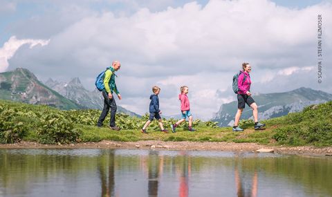 Eine vierköpfige Familie wandert auf einem malerischen, grasbewachsenen Pfad mit einem ruhigen Teich im Vordergrund. Im Hintergrund sind Berge unter einem teilweise bewölkten Himmel zu sehen. Jedes Mitglied trägt farbenfrohe Wanderkleidung und einen Rucksack.