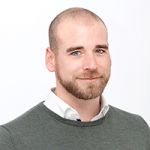 Ein Mann mit rasiertem Kopf und Bart lächelt leicht. Er trägt ein weißes Hemd unter einem grauen Pullover. Der Hintergrund ist schlicht weiß.