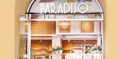 Ein Restauranteingang mit dem Schild „Paradiso“ in fetten weißen Buchstaben über der Tür. Im Inneren sorgen warmes Licht, Backsteinwände und Pflanzen für eine gemütliche Atmosphäre. Über der Theke sind Menütafeln sichtbar.