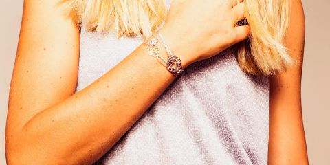 Nahaufnahme einer Person mit langem, hellem Haar, die ein graues ärmelloses Oberteil trägt. Sie zeigt ein silbernes Armband mit komplizierten Mustern an ihrem Handgelenk und ihre Hand berührt sanft ihren Hals.