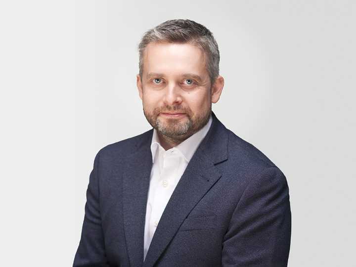 Ein Mann mit kurzen grauen Haaren und Bart trägt ein dunkles Jackett und ein weißes Hemd. Er sitzt vor einem schlichten weißen Hintergrund und blickt mit neutralem Gesichtsausdruck direkt in die Kamera.