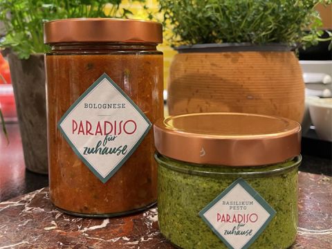 Auf einer Marmorplatte stehen zwei Einmachgläser mit der Aufschrift „Paradiso für zuhause“. Das größere Glas enthält Bolognesesauce, das kleinere Basilikumpesto. Im Hintergrund sind Topfpflanzen zu sehen.
