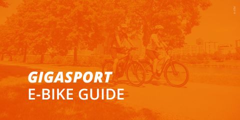 Zwei Personen fahren mit E-Bikes auf einem Weg mit Bäumen und Stadtgebäuden im Hintergrund. Das Bild ist mit einem orangefarbenen Farbton überlagert. Der Text lautet: „Gigasport E-Bike Guide.