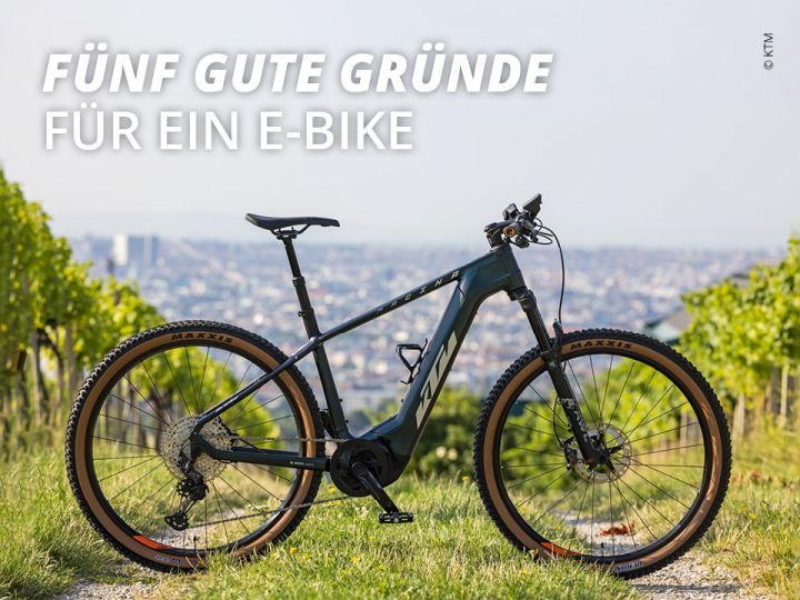 Ein schwarzes E-Bike steht auf einem Grasweg in einem Weinberg. Im Hintergrund ist eine Stadtlandschaft unter einem strahlend blauen Himmel zu sehen. Der Text auf dem Bild lautet „Fünf gute Gründe für ein E-Bike“.