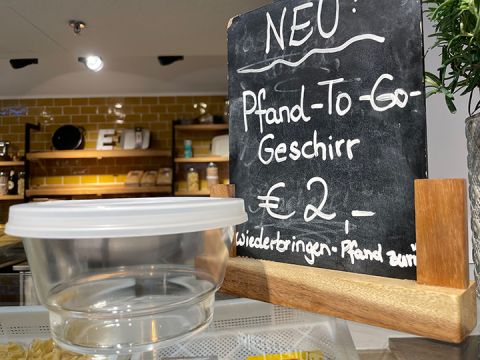 Ein Thekenaufsteller mit einem Tafelschild mit der Aufschrift „NEU: Pfand-To-Go Geschirr 2 €“. Unter dem Schild steht ein durchsichtiger, wiederverwendbarer Lebensmittelbehälter mit weißem Deckel. Im Hintergrund sind Regale mit verschiedenen Artikeln zu sehen.