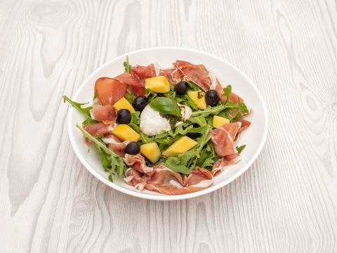 Auf einem weißen Teller auf einer Holzoberfläche liegt ein Salat mit Rucola, Prosciuttoscheiben, Mangowürfeln, schwarzen Oliven, einem Klecks Käse und garniert mit einem Basilikumblatt.