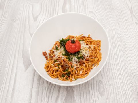 Eine Schüssel Spaghetti, belegt mit geriebenem Käse, gehackten Kräutern und einer gerösteten Kirschtomate. Das Gericht steht auf einer hellen Holzoberfläche.