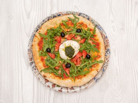 Eine Pizza mit Rucola, Tomatenwürfeln, schwarzen Oliven und einer großen Kugel Burrata-Käse in der Mitte, beträufelt mit Pesto, serviert auf einem dekorativen Teller vor einem hellen Holzhintergrund.