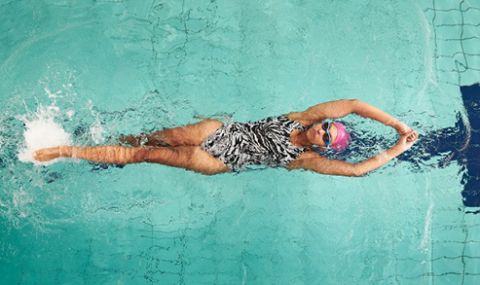 Ein Schwimmer in einem gemusterten Badeanzug und einer rosa Badekappe schwimmt in einem klaren, blauen Pool Rückenschwimmen. Die Arme sind über dem Kopf ausgestreckt und das Wasser kräuselt sich um ihn herum.