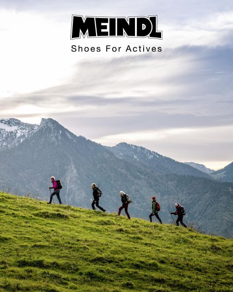 Eine Gruppe Wanderer mit Rucksäcken geht in einer Reihe einen grasbewachsenen Hügel hinauf. Im Hintergrund sind schneebedeckte Berge und ein bewölkter Himmel zu sehen. Oben auf dem Bild steht der Text „MEINDL Shoes For Actives“.