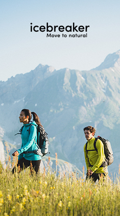 Zwei Wanderer mit Rucksäcken durchqueren eine Wiese mit gelben Blumen vor der Kulisse majestätischer Berge. Der Text lautet „Eisbrecher – Bewegen Sie sich in die Natur“. Die Szene vermittelt ein Gefühl von Abenteuer und Verbundenheit mit der Natur.