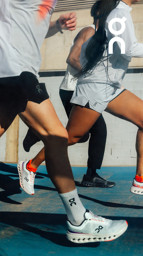 Eine Gruppe von Menschen läuft in Sportkleidung auf einer blauen Laufbahn. Sichtbares Branding auf Schuhen und Kleidung. Teilansicht, fokussiert auf die Beine in Bewegung. Helle Außenkulisse mit Schatten auf dem Boden.