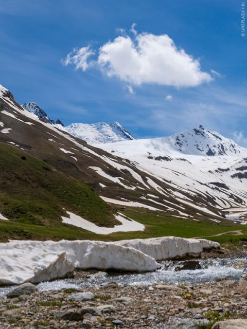 Eine malerische Berglandschaft mit schneebedeckten Gipfeln unter einem klaren blauen Himmel. Ein kleiner Bach fließt durch den Vordergrund, flankiert von Schneefeldern und einem grasbewachsenen Hang. Darüber schwebt eine einzelne flauschige Wolke.