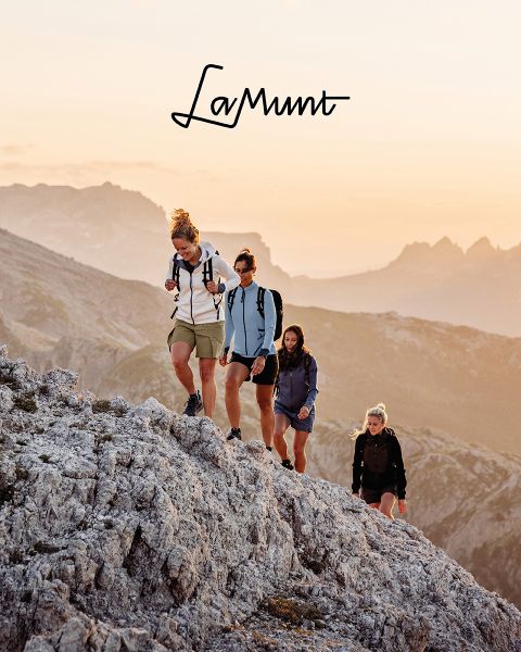 Drei Personen in Winterkleidung fahren unter klarem Himmel Ski durch eine verschneite Berglandschaft. Der Text lautet: „LaMunt MOUNTAIN ME-TIME“. Im Hintergrund sind schneebedeckte Gipfel zu sehen.