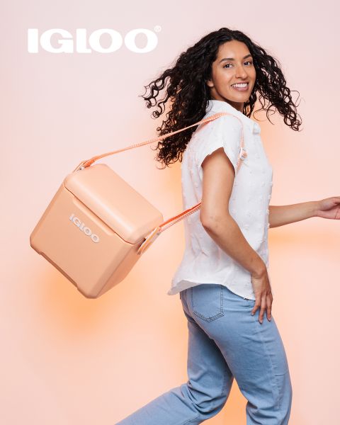 Eine lächelnde Frau mit lockigem Haar trägt eine pfirsichfarbene Igloo-Kühlbox an einem Seilgurt über der Schulter. Sie trägt eine weiße Kurzarmbluse und hellblaue Jeans und steht vor einem hellpfirsichfarbenen Hintergrund.