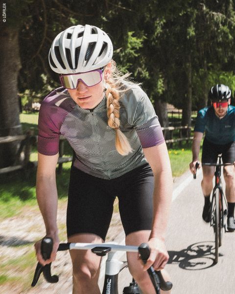 Eine Frau mit blondem Zopf, Fahrradhelm, Sonnenbrille und Radtrikot fährt Rennrad, gefolgt von einem männlichen Radfahrer auf einer sonnigen, von Bäumen und einem Holzzaun gesäumten Straße.