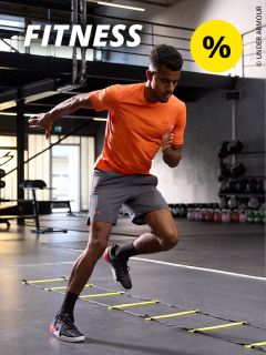 Ein Mann in orangefarbenem Hemd und grauen Shorts macht in einem Fitnessstudio Koordinationsübungen mit der Leiter. In der oberen linken Ecke sind das Wort „FITNESS“ und ein gelbes Prozentzeichen eingeblendet. Im Hintergrund sind Kettlebells und Fitnessgeräte zu sehen.