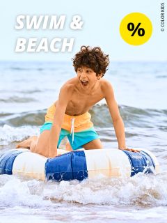 Ein lächelnder Junge in einer gelb-türkisen Badehose reitet auf einem blau-weißen aufblasbaren Ring im seichten Meerwasser. Der Text „SWIM & BEACH“ und ein gelbes Rabattsymbol sind zu sehen.