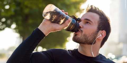 Ein Mann in einem schwarzen langärmeligen Hemd und mit Kopfhörern trinkt Wasser aus einer Sportflasche im Freien, mit Bäumen und Sonnenlicht im Hintergrund.