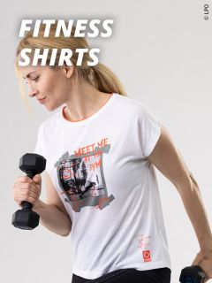 Eine Frau in einem weißen grafischen T-Shirt hebt eine Hantel mit ihrem rechten Arm. Der Text auf dem Bild lautet "FITNESS SHIRTS". Sie scheint konzentriert zu sein und steht vor einem hellgrauen Hintergrund.