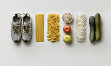 Ein Paar Laufschuhe neben ordentlich angeordneten Lebensmitteln: ungekochte Spaghetti, Penne-Nudeln, Haferflocken, zwei Äpfel, weißer Reis und zwei Zucchini, alle aufgereiht auf einem weißen Hintergrund.
