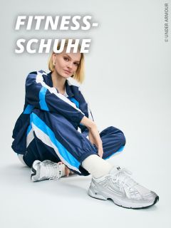 Eine Person in einem blau-weißen Trainingsanzug sitzt auf dem Boden, lächelt und trägt weiße Under Armour Turnschuhe. In der oberen linken Ecke steht in Fettschrift "FITNESS-SCHUHE".