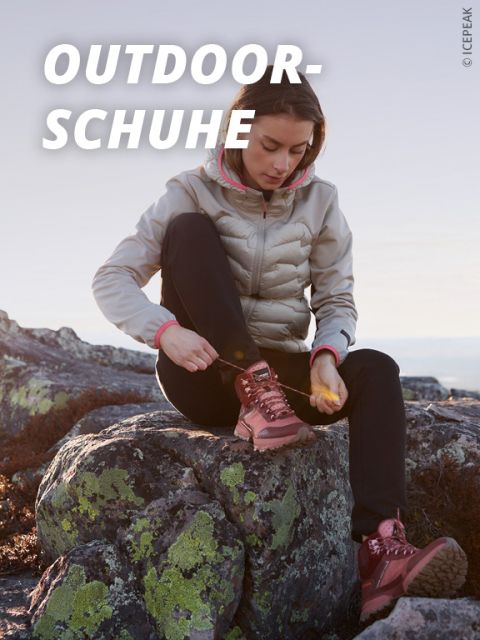 Eine Frau sitzt auf einer felsigen Fläche im Freien und bindet die Schnürsenkel ihrer rosa Wanderschuhe. Sie trägt eine leichte Jacke und eine schwarze Hose. Das Wort "OUTDOORSCHUHE" erscheint in fetter weißer Schrift auf dem Bild.