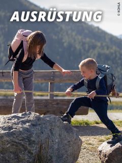 Eine Frau hilft einem Jungen beim Klettern über Felsen an einem Seil im Freien. Beide tragen Rucksäcke, und im Hintergrund sind Berge zu sehen. Oben ist das Wort "Ausrüstung" zu lesen.