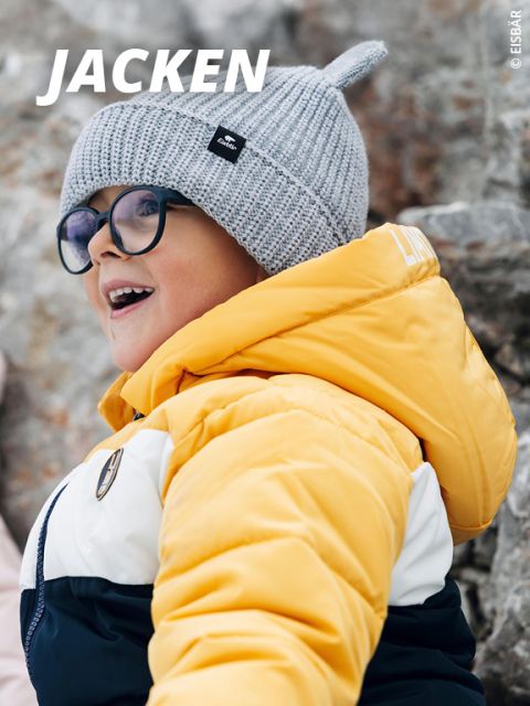 Lächelndes Kind in gelb-weißer Winterjacke und grauer Strickmütze mit Ohrenklappen, das eine Brille trägt, steht im Freien in einer verschneiten Umgebung. Das Wort "JACKEN" steht in fetter weißer Schrift oben links.