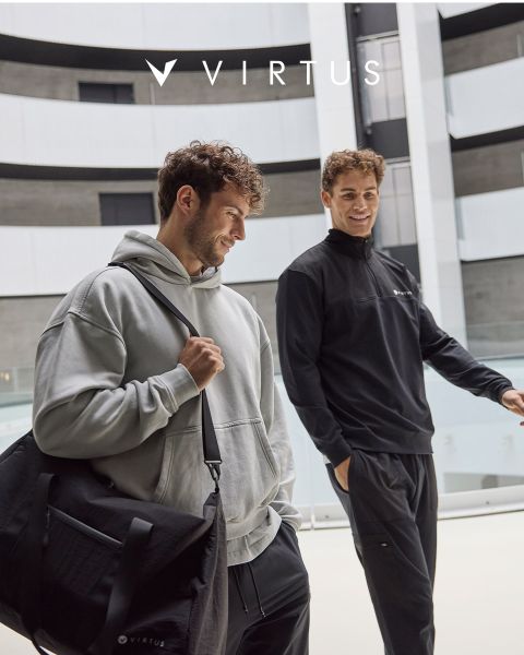 Zwei Männer in Sportkleidung von Virtus, der eine in einem hellgrauen Kapuzenpulli mit schwarzem Seesack, der andere in einer schwarzen Jacke, lächeln und gehen zusammen in einer modernen Halle spazieren. Das Virtus-Logo ist über ihnen zu sehen.