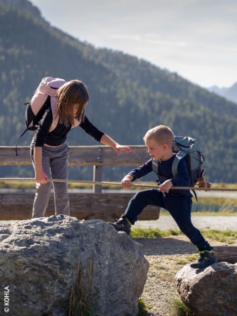 Drei Kinder laufen in einem Wald an einem umgestürzten Baum entlang. Sie sind in warme, farbenfrohe Kleidung gekleidet. Der Hintergrund ist voller grüner Bäume. Der Text lautet: „NACHHALTIGES FÜR KINDER.
