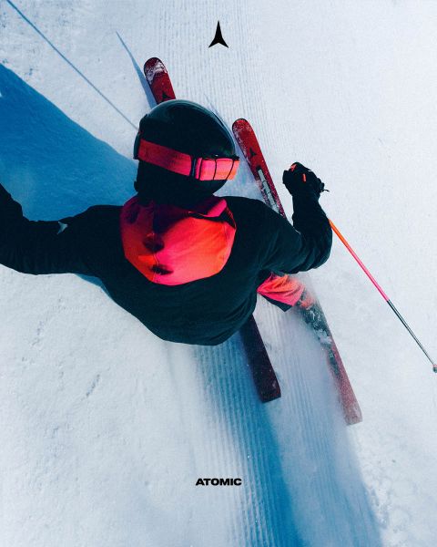Ein Skifahrer in einem schwarz-roten Outfit und Helm fährt auf präpariertem Schnee bergab, von oben gesehen. Das Atomic-Logo ist oben zu sehen und der Atomic-Markenname steht unten im Bild.