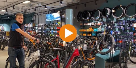 Ein Fahrradladen mit einer Person neben einem E-Trekkingrad. An den Wänden des Ladens sind verschiedene Fahrräder, Zubehör und Reifen ausgestellt. Der Text „E-TREKKINGBIKE TIPP“ überlagert das Bild. In der Mitte befindet sich ein orangefarbener Play-Button.