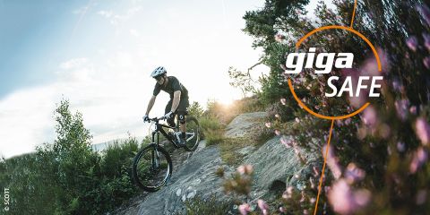 Ein Radfahrer mit Helm fährt einen steinigen Weg entlang, der von Bäumen und Blumen umgeben ist. Der Himmel ist teilweise bewölkt. Der Text lautet „Sicherheit beim Radfahren“ und „giga SAFE“ mit einem kreisförmigen Design.