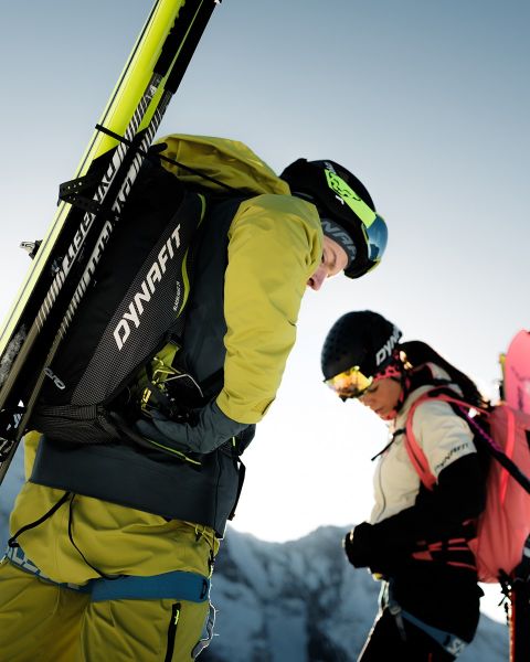 Zwei Personen laufen auf einem Bergpfad mit einer Stadt und einem Fluss im Hintergrund. Einer trägt ein schwarzes Shirt und Shorts, der andere ein blaues Tanktop und Shorts. Das Dynafit-Logo befindet sich oben im Bild.