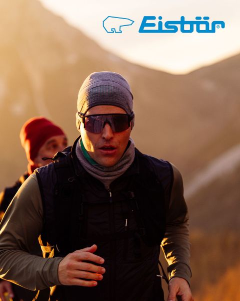 Eine Frau und ein Mann lächeln beim Skifahren im verschneiten Gelände. Sie trägt eine rote Jacke und eine weiße Mütze und hält Skistöcke in der Hand, er eine beige Jacke und eine grüne Mütze. Über ihnen prangt das „Eisbär“-Logo.