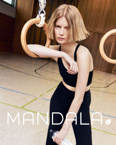 Frau mit Mütze und schwarzem Tanktop blickt direkt in die Kamera. Ihr Haar ist wellig und offen. Das Bild ist schwarz-weiß und trägt unten in großen Buchstaben das Wort „MANDALA“.