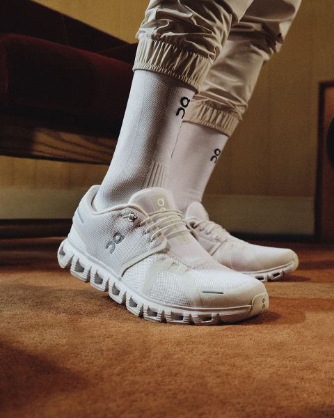 Nahaufnahme einer Person, die weiße On-Running-Turnschuhe und passende weiße On-Socken trägt und auf einem orangefarbenen Teppich mit beiger Hose steht. Im Hintergrund sind eine Holzwand und ein kastanienbraunes Möbelstück zu sehen.