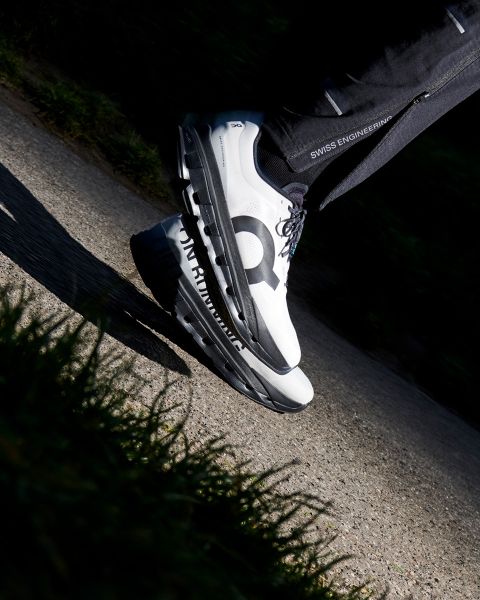 Eine Person mit weißen und schwarzen Laufschuhen mit der Aufschrift "Swiss Engineering" auf der Hose steht auf einem sonnenbeschienenen Bürgersteig und wirft einen Schatten. Der Schuh trägt ein auffälliges schwarzes "Q" an der Seite.